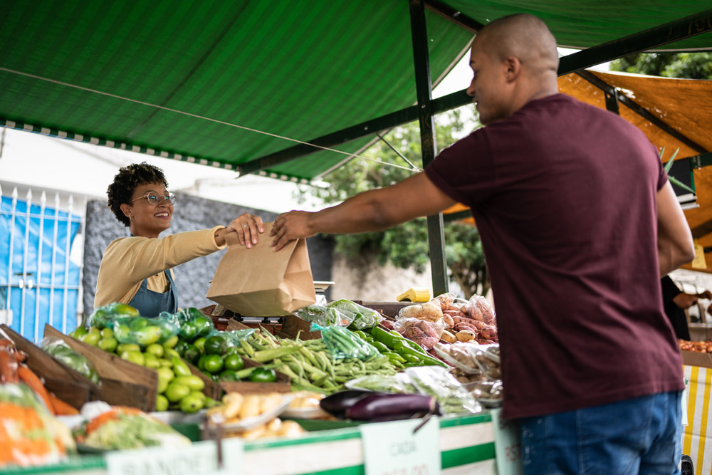 A woman hands a man a brown paper bag from a farmers market vegetable stand