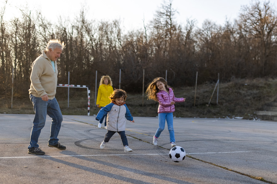 An older man plays soccer or kickball with young children on a paved blacktop surface at a park, while an older women in a yellow coat watches in the background