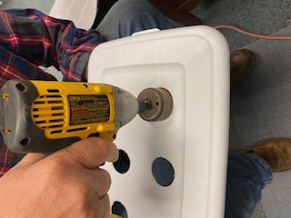 A hand-held drill bores holes into a plastic lid.