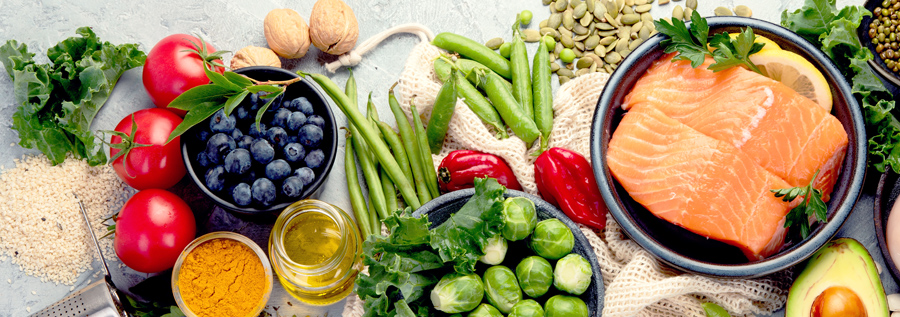 A tabletop is covered with nutrient-rich foods such as green and red vegetables, salmon, beans, an avocado, and blueberries