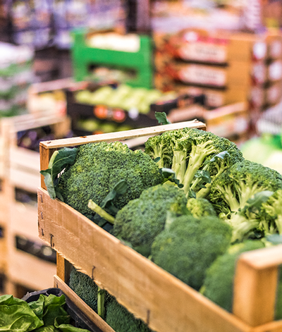 A crate of broccoli sits in a market setting, showing fruits and vegetables in other packages.