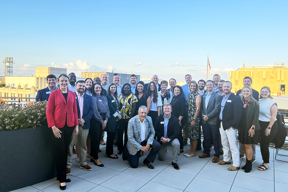 Group of adults posing on a rooftop with a cityscape and blue sky in the background, dressed in business and professional attire.