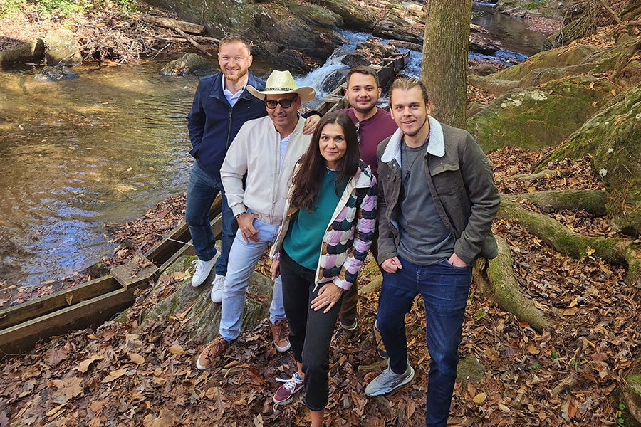 Five people posing by a wooded stream with fallen leaves, wearing casual clothes. A small waterfall is in the background.