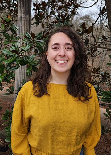 A young woman with brown hair and a yellow sweater smiles at the camera with foliage in the background