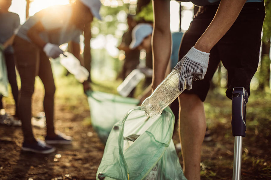 A volunteer places a clear, empty plastic bottle into a partially filled, translucent green trash bag. Other volunteers are visible in the background, also picking up trash; the sun is shining through the trees.