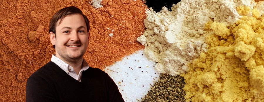 A headshot of a smiling man with brown hair is superimposed on a background of dried, powdered spices.