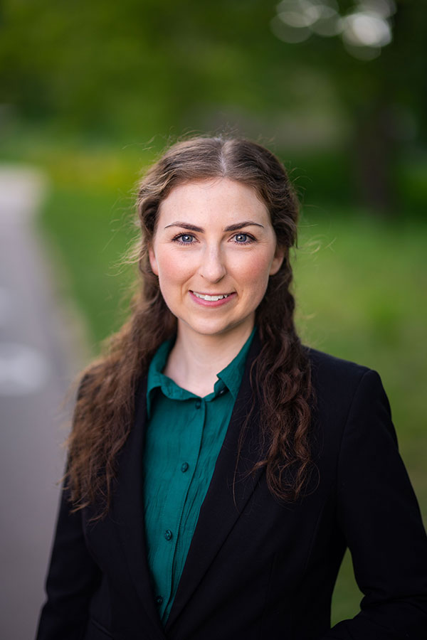 A young woman with long wavy brown hair, wearing a dark blazer over a teal green button-up shirt, stands outdoors on a paved path with blurred greenery in the background. She is smiling and looking directly at the camera in a professional headshot.