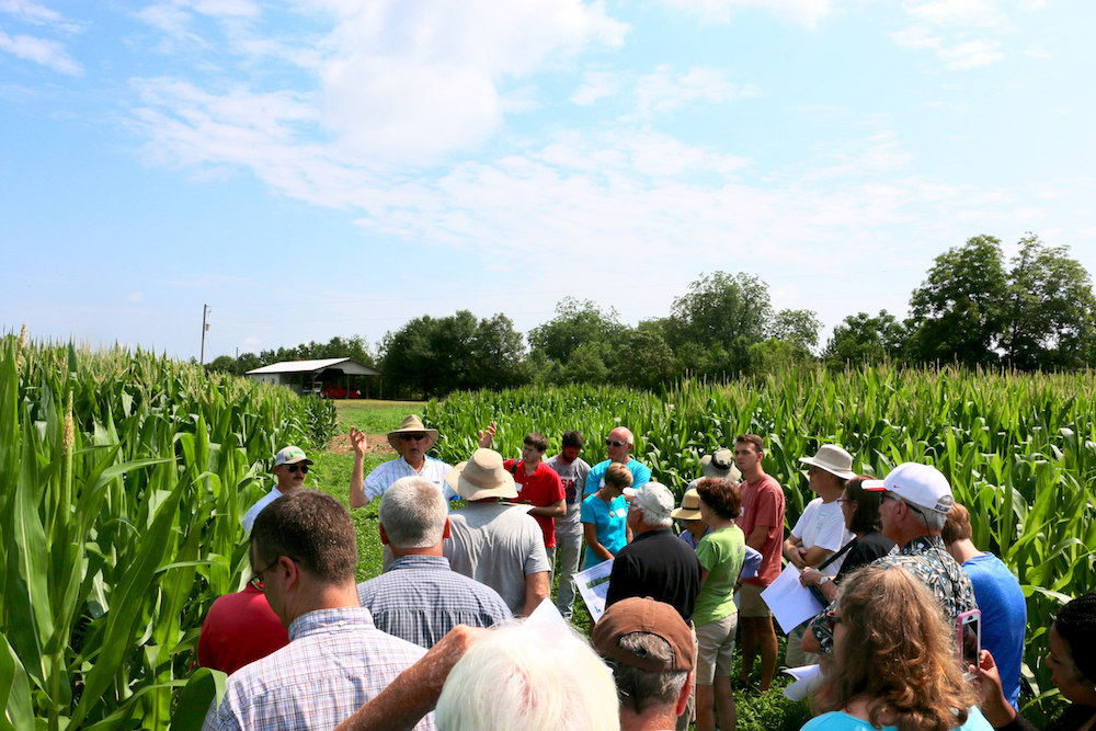 Twenty-three mixed gender and mixed race attendees listen to a white, male speaker in a corn field during a farm tour.