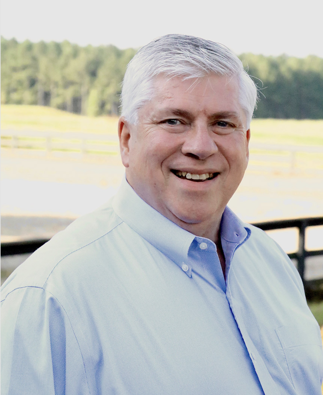 Francis Fluharty, a white male wearing a blue button-down shirt, smiles in front of a pasture.