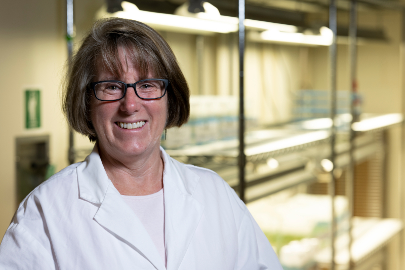 Robin Buell smiles in her lab, wearing a white lab coat and glasses and standing in front of a set of shelves