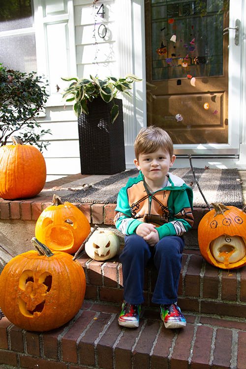 Boy sits on a front stoop with a variety of carved pumpkins