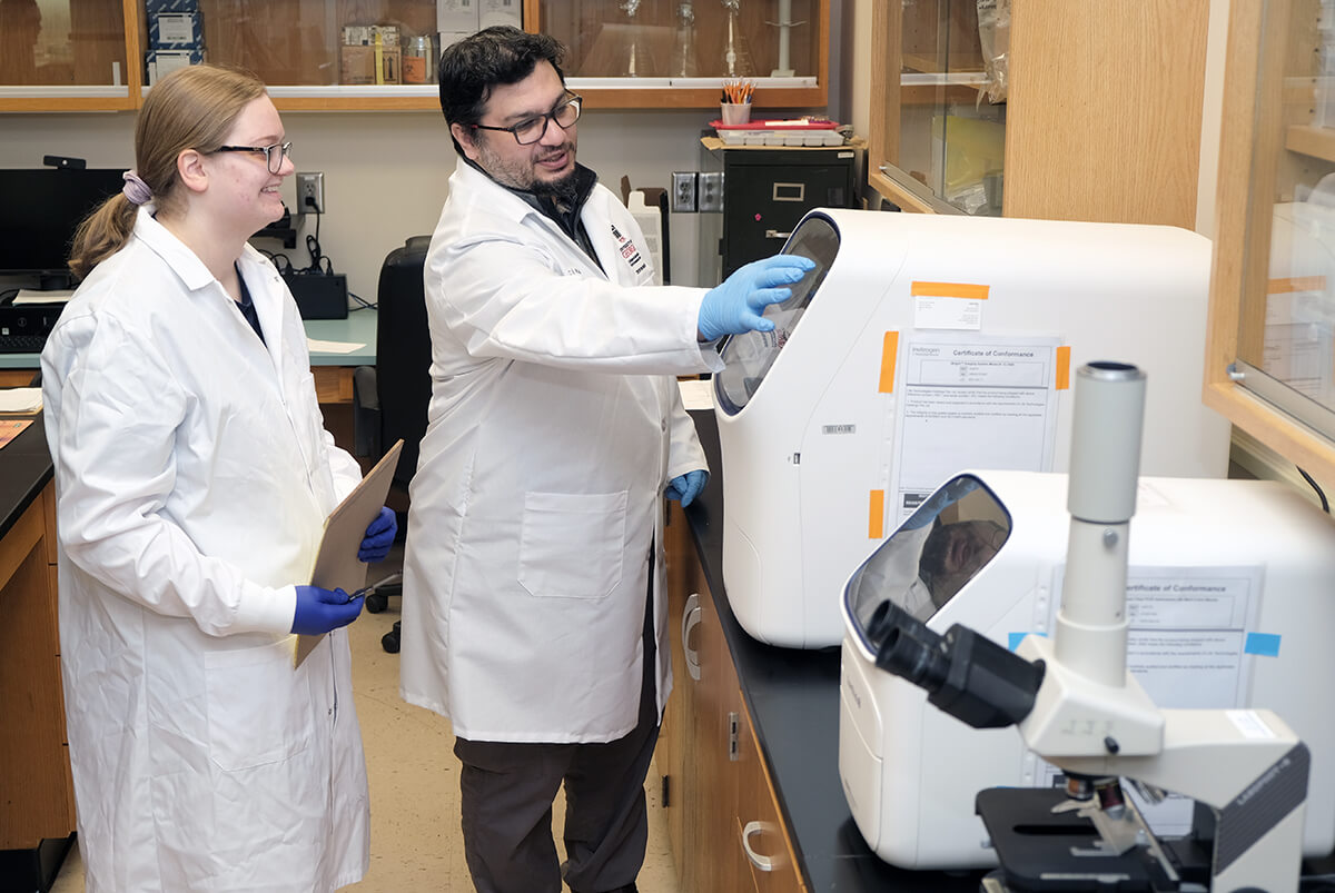 Miranda Barr (left) and Issmat Kassem work in Kassem's research lab wearing white lab coats with lab equipment on the right