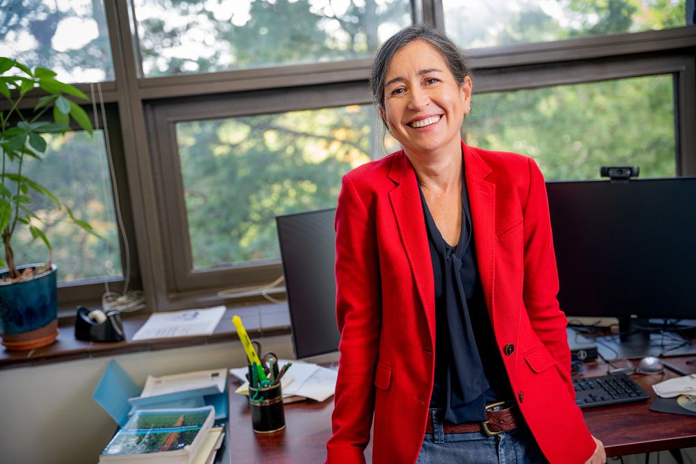 Economist Susana Ferreira sits on her desk in her office while smiling and wearing a bright red blazer