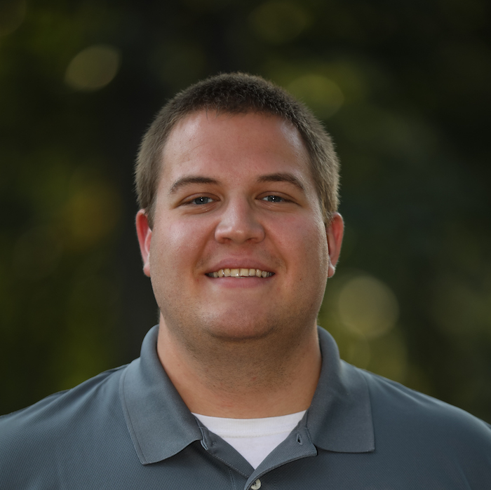 Matthew Holton smiles in front of greenery while wearing a gray collared shirt