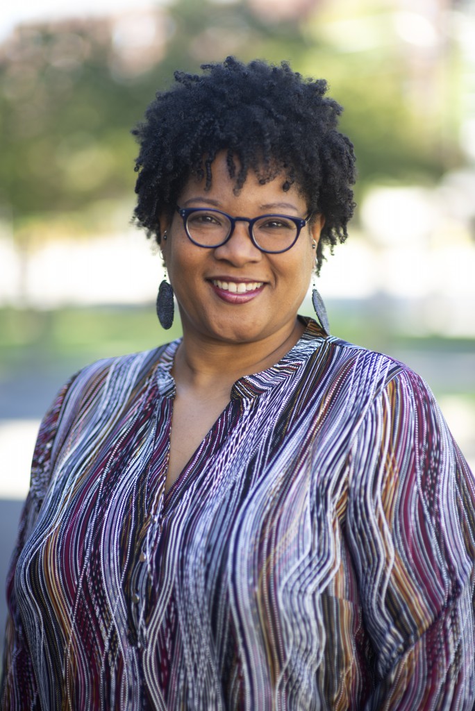 Monica M. White smiles in her professional headshot, wearing a purple top, acetate-rimmed glasses, and earrings