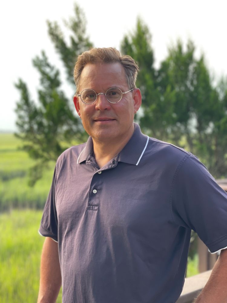 Jim Gratzek, a white male, smiles while wearing a blue polo shirt in an outdoor setting with trees and grass behind him