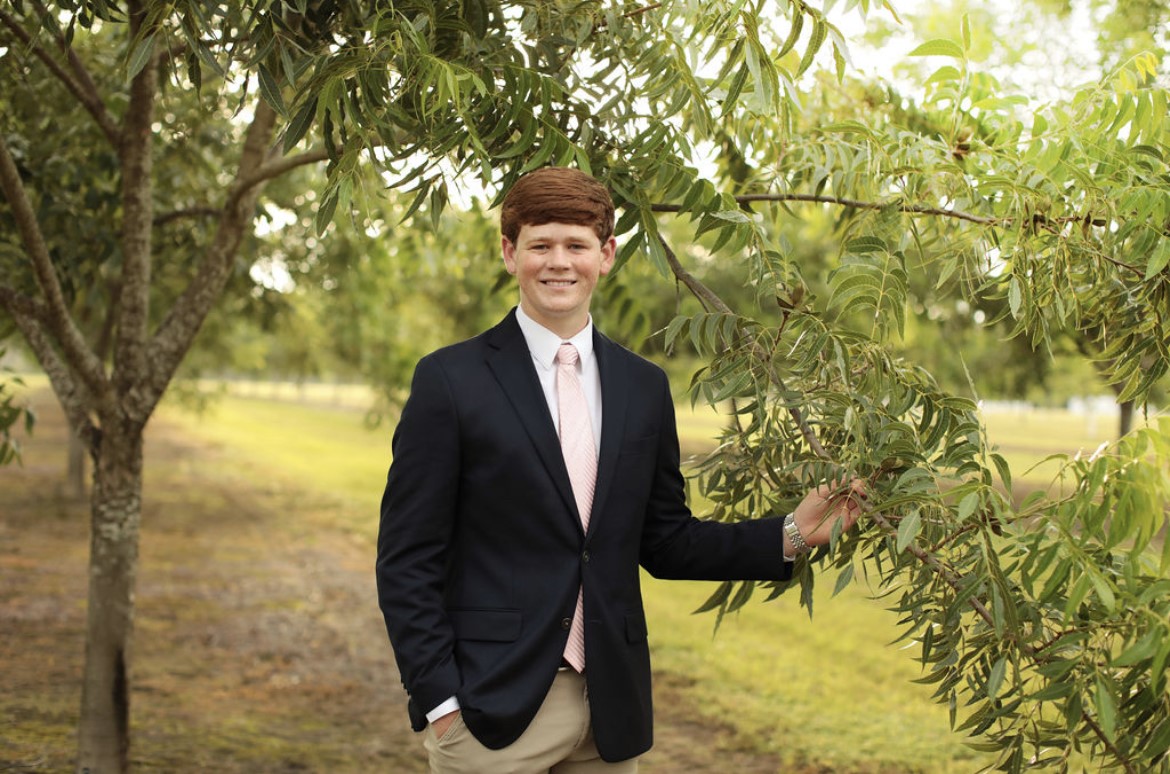 Young, white male with red hair stands in a pecan grove wearing khaki pants, a white shirt and black suit jacket. He right hand is in his pocket and his left is holding a pecan tree branch.