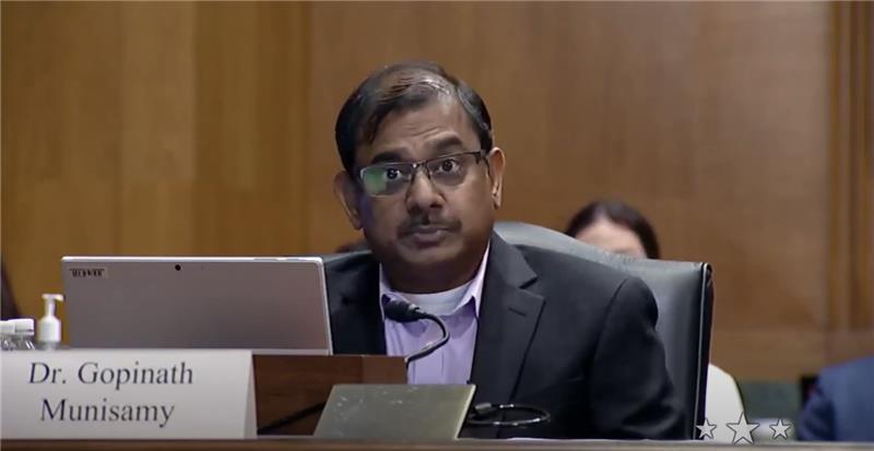 Gopinath Munisamy, a male with medium brown skin and black hair, wears a lavender dress shirt and black blazer while sitting at a desk during a Senate Hearing.