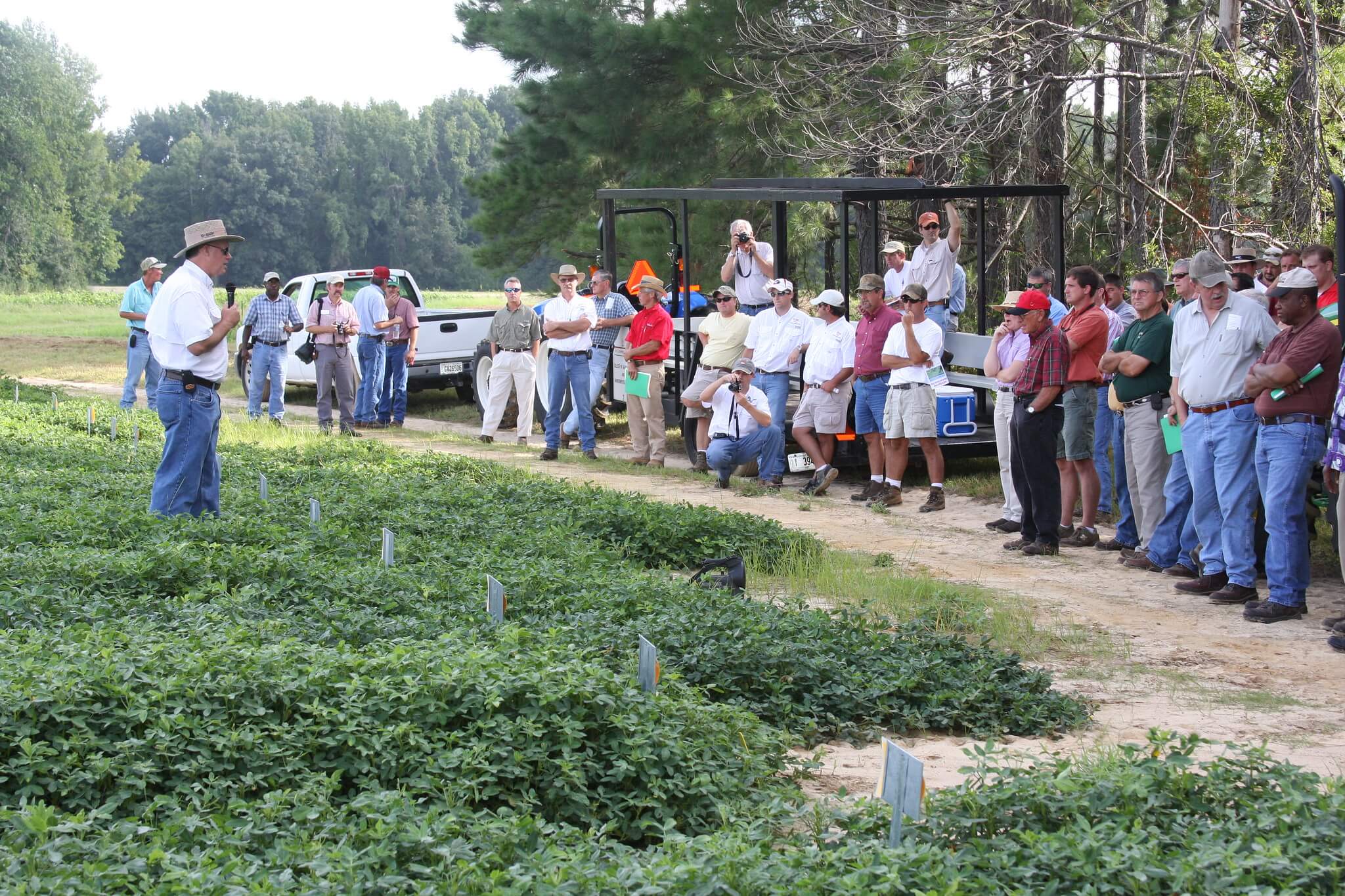 A faculty member discusses field research in front of a crowd, mostly dressed in sun hats and jeans on the research farm.