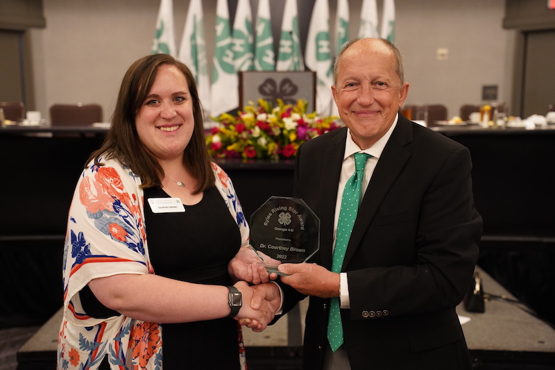 National 4-H Council Director Roger C. “Bo” Ryles presents Extension Specialist Courtney Brown with the Ryles Rising Star Award in front of the center lectern decorated with 4-H flags and flowers.