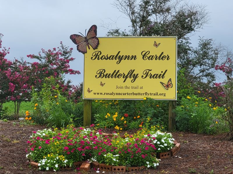 Yellow sign with name and website for Rosalynn Carter Butterfly Trail in a field of flowers