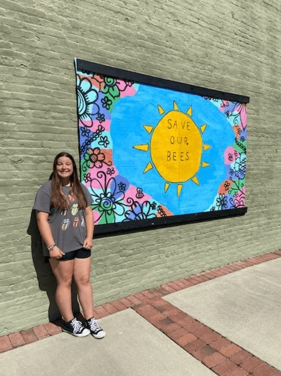 New pollinator census grand marshal Mia Burnett, a member of Georgia 4-H, stands in front of her bee mural outside, which says "save our bees" within a sun illustration.