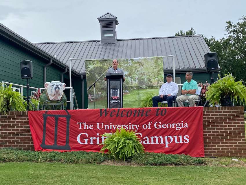 Clint Waltz speaks at a lectern on an outdoor stage, which is decorated with a banner that reads, "Welcome to the University of Georgia Griffin Campus." Two men, David Buntin and Doug Hollberg, are seated on the right side of the stage.