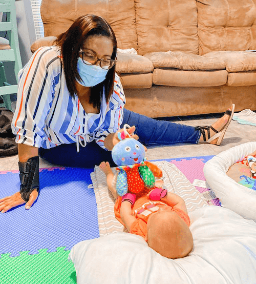 Home visitor Rafaelina Sanchez, wearing a mask and wrist brace, works with a baby on a floor mat of a client's home.