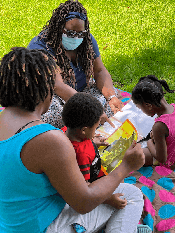 Wearing a mask and sunglasses, home visitor Ashley Maddox works with a mom and two young children on a blanket in the grass, where all are occupied in reading colorful children's stories.