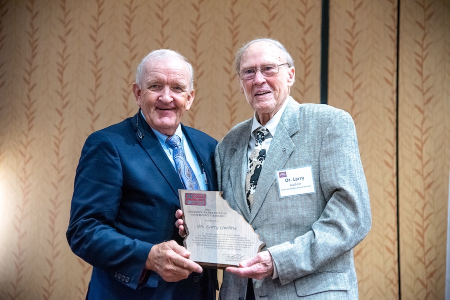 A white male wearing a dark blue suit and tie stands next to a white male in a grey suit, tie and glasses. The men are holding a wooden award cut in the shape of Georgia.