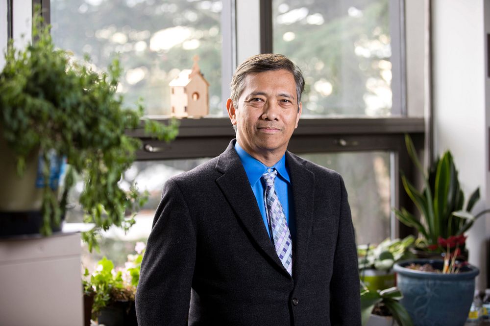 Environmental portrait of UGA Professor Cesar Escalante, a smiling man with light brown skin and short, salt-and-pepper hair wearing a black suit coat, medium blue dress shirt, and a tie with squares in multiple shades of blue. He is surrounded by house plants lit by a wall of windows behind him.