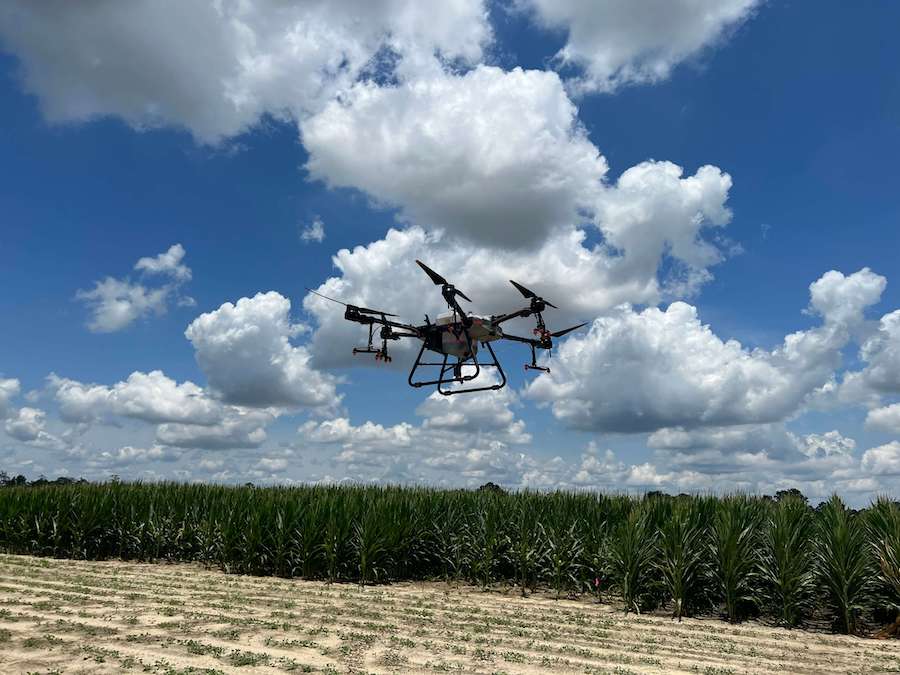 A sprayer drone flies above a UGA research field on a partly cloudy day