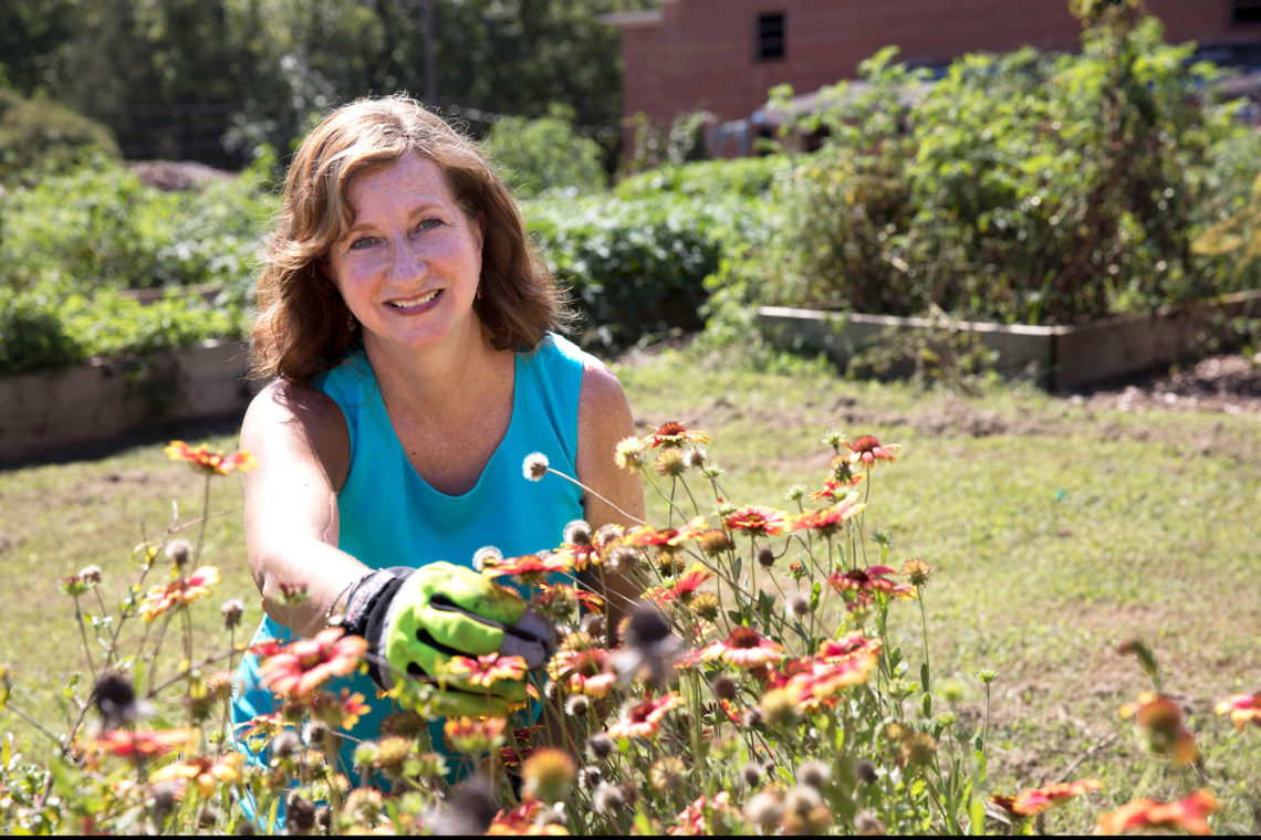 Becky Griffin, Extension school and community garden coordinator wearing a blue tank top and gardening gloves, tends to flowers in a raised bed garden in the sunshine.