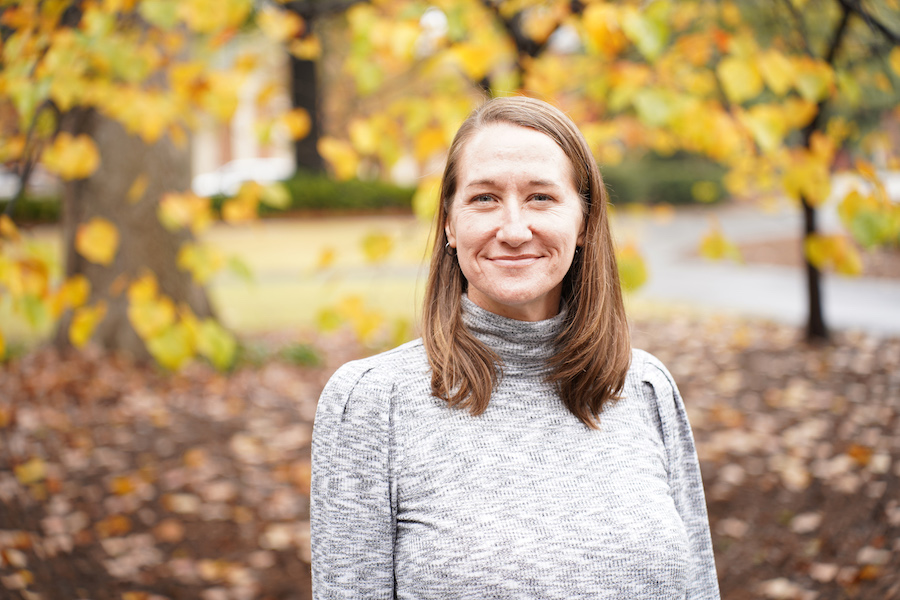 Portrait of Melanie Biersmith, wearing a gray heather turtleneck sweater against a backdrop of trees with colorful fall leaves. 