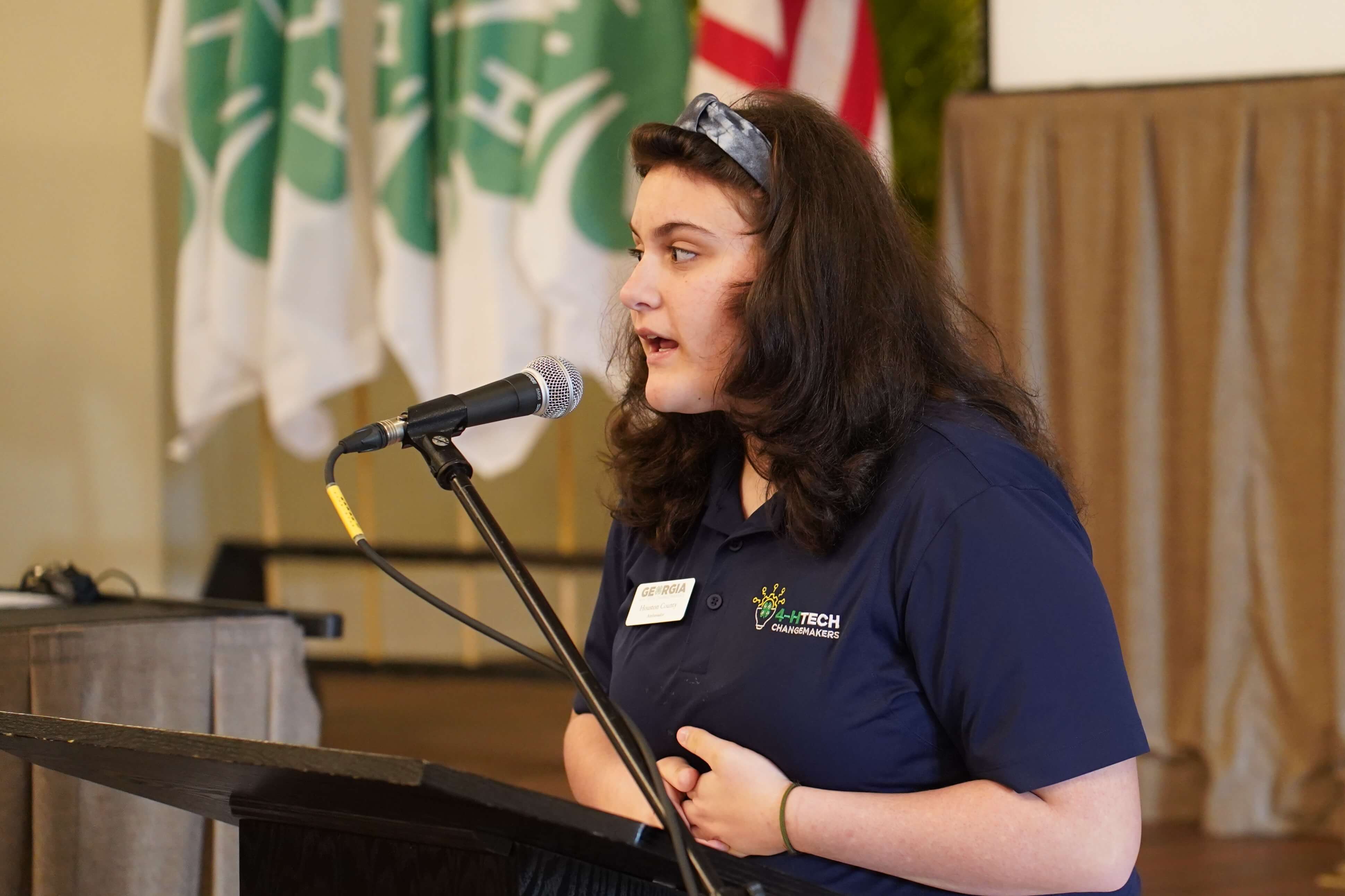 Houston County 4-H Tech Changemaker Leilani Priest-Akens addresses 130 university leaders at a regional conference while standing at a lectern and speaking into a microphone. She wears a collared shirt with the 4-H Tech Changemakers logo and flags representing the U.S. and 4-H are visible in the background.