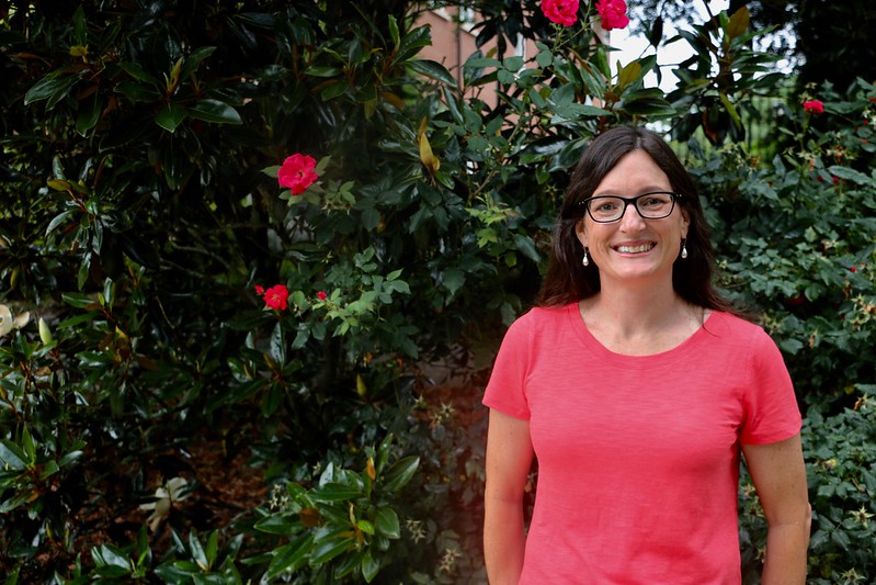 Woman with glasses and short sleeved, pink shirt stands in front of lush greenery with pink flowers.