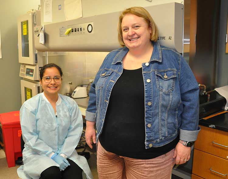Martha Sanchez is seated next to a laboratory fume hood as Associate Professor Faith Critzer stands next to her. 