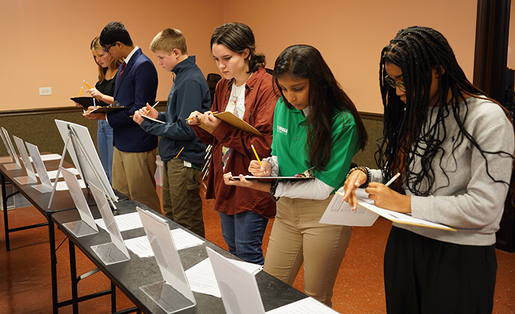 Six 4-H students, including students of a variety of genders and ethnicities, stand in front of a judging table to assess consumer situations and use critical thinking skills to rank the best options for given consumer needs.
