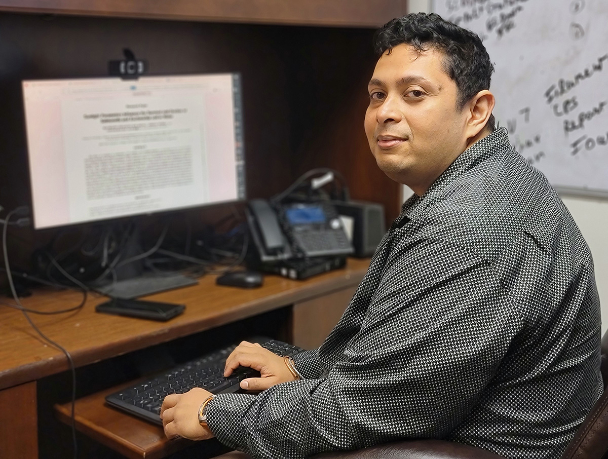 Professor Dev Kumar looks back at the camera while sitting at his desktop computer, with a whiteboard to his right.
