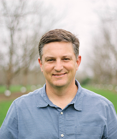 A white male in a blue shirt stands in a pecan orchard.