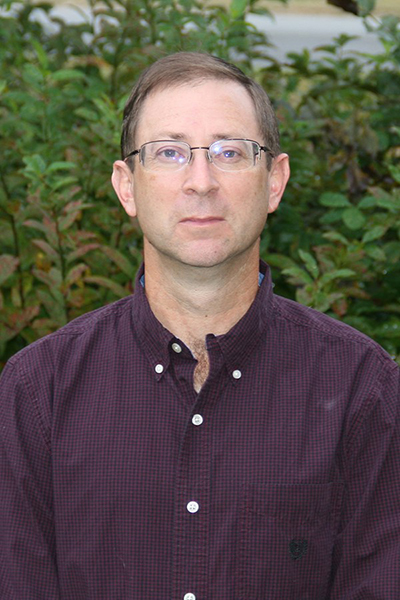 A white male wearing glasses and a burgundy shirt stands in front of greenery