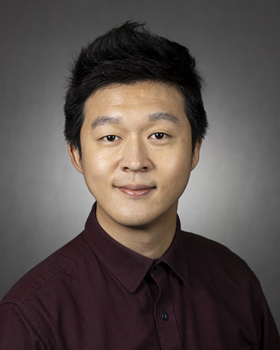 An Asian male wearing a burgundy shirt stands in front of a grey background