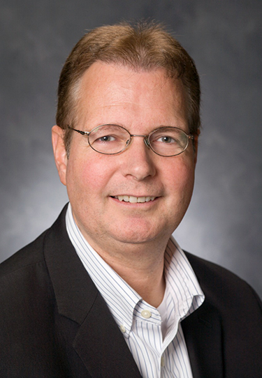 A white male wearing glasses, a grey and white striped shirt and black jacket stands in front of a grey background.