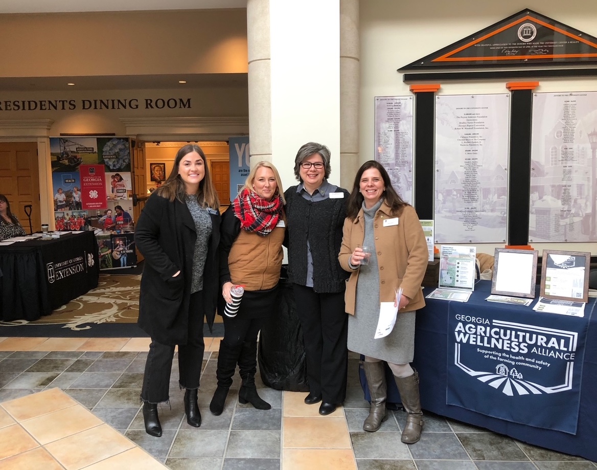 Four women gather before the Farm Stress Summit, standing in front of materials branded for the Georgia Agricultural Wellness Alliance and UGA Extension