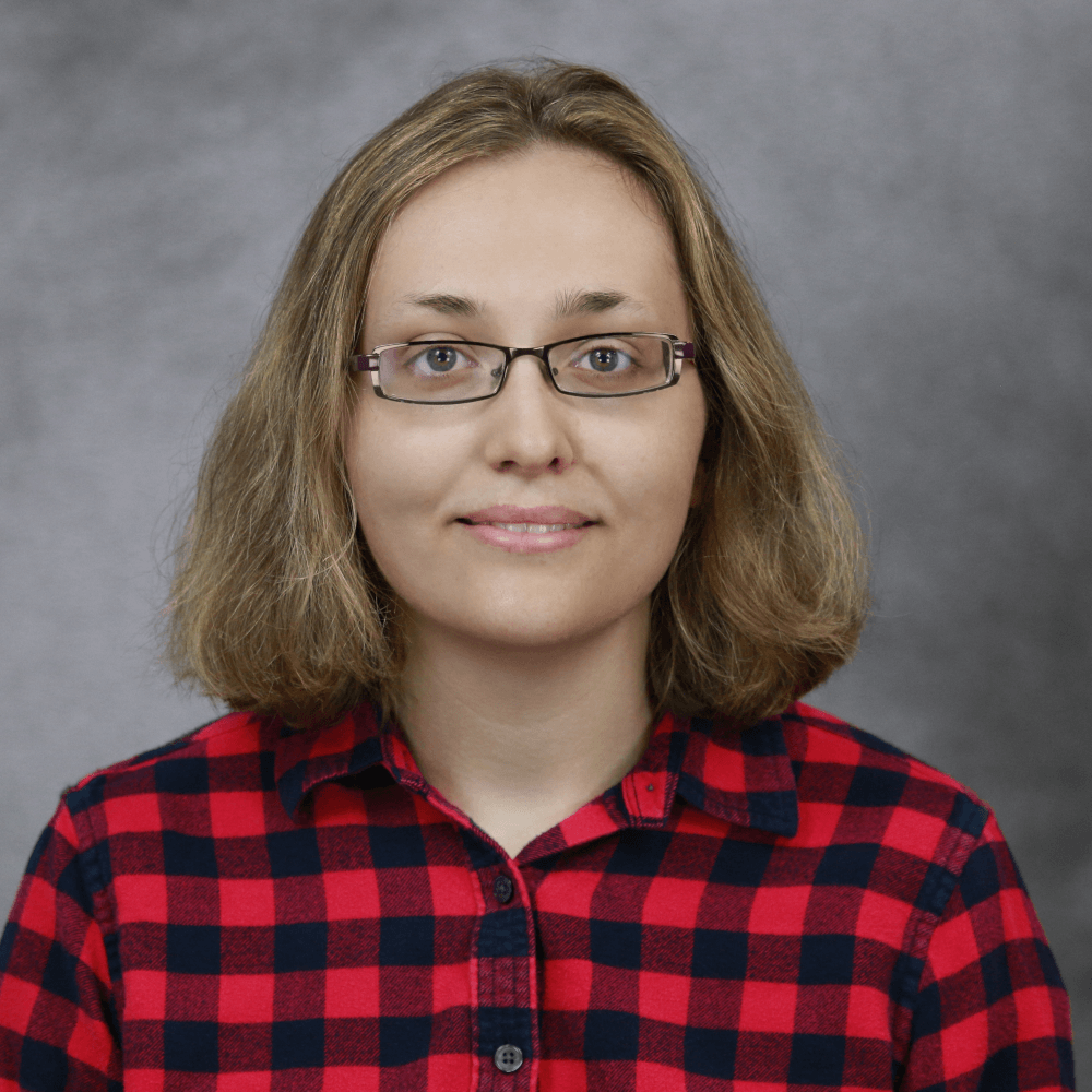 A person with shoulder-length, light brown hair looks directly at the camera, wearing a red and black plaid shirt and glasses.