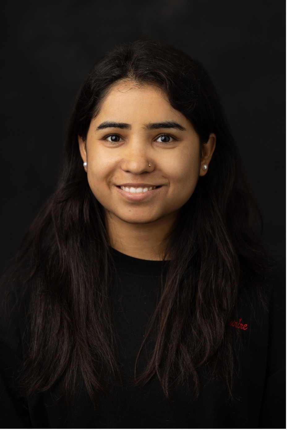 A person with long brown hair wearing a black shirt looks directly at the camera, smiling.