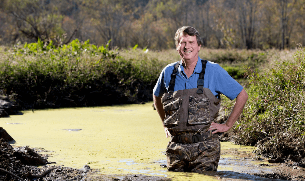 Darold Batzer, wearing waders and a blue collared shirt, stands in a wetland.