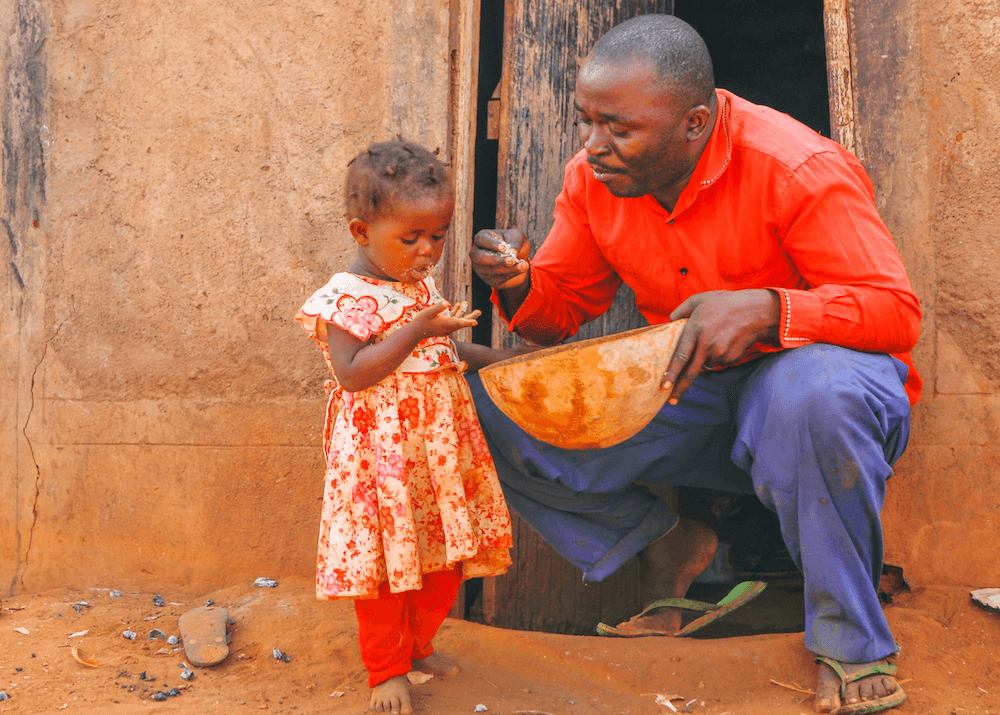 In a doorway, a man feeds a young girl finger millet
