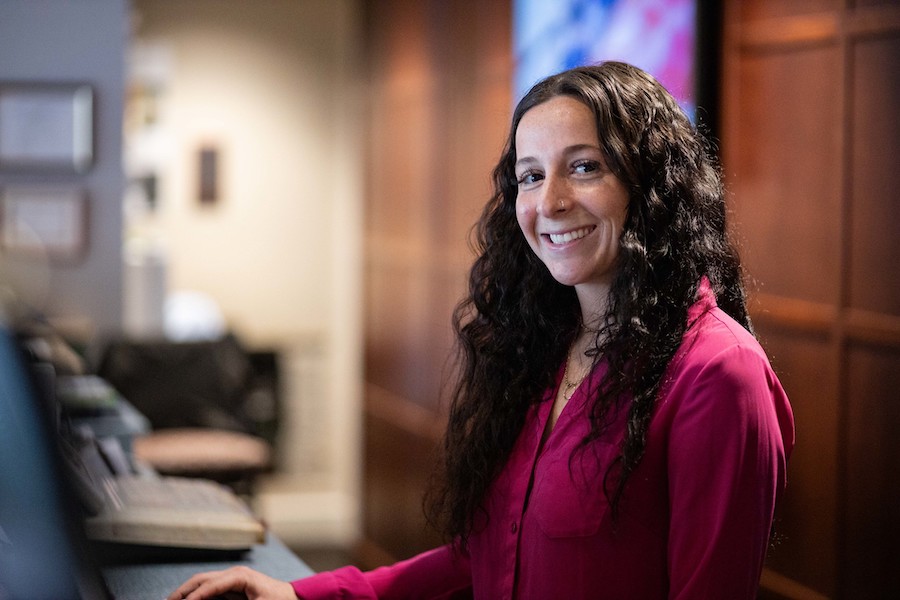 A woman with curly brown hair smiles at camera at a hotel reception desk