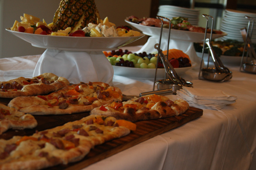 A buffet at an event with pizza, fruit and cheese.